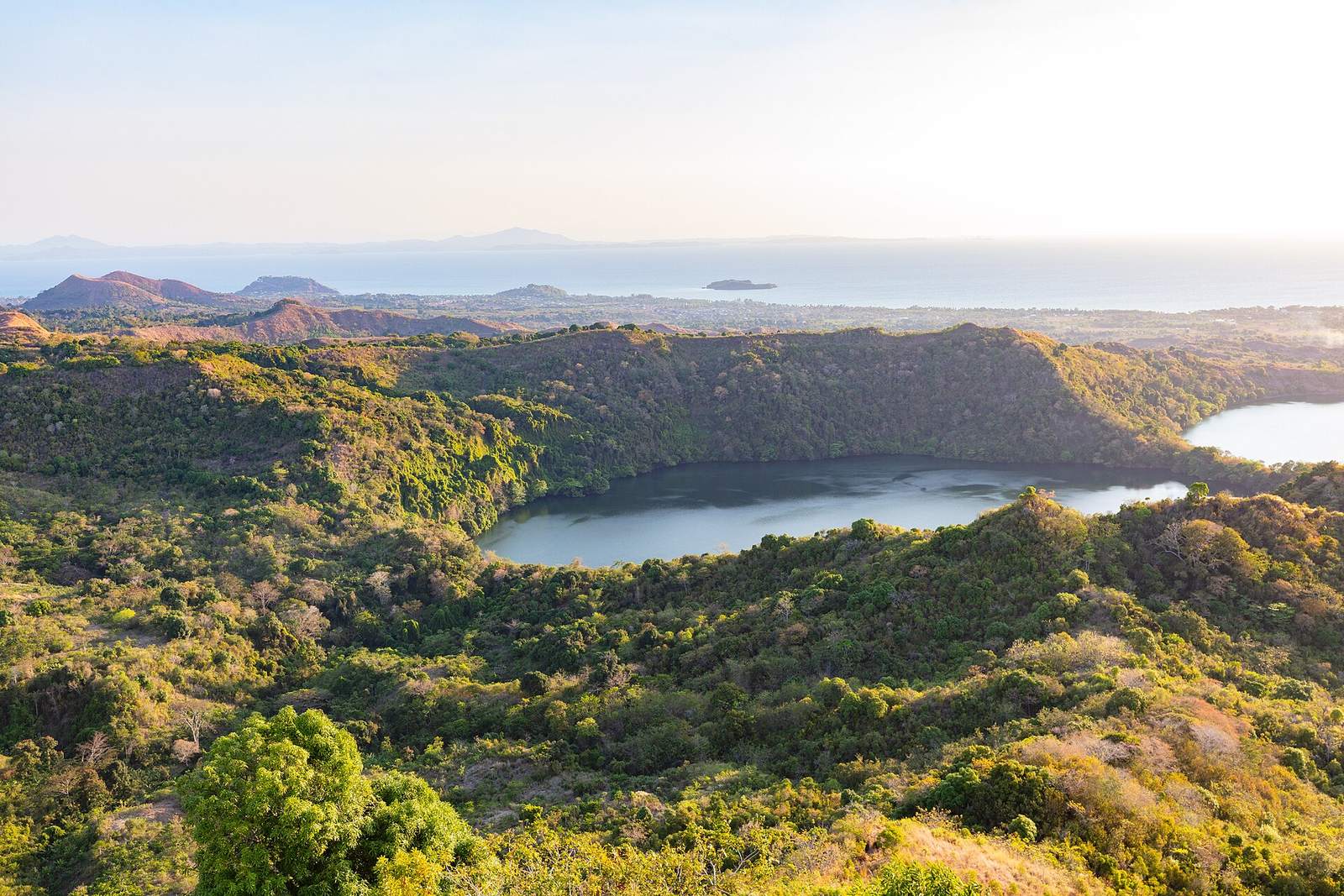 A forest-ringed crater lake on Nosy Be with the sea beyond